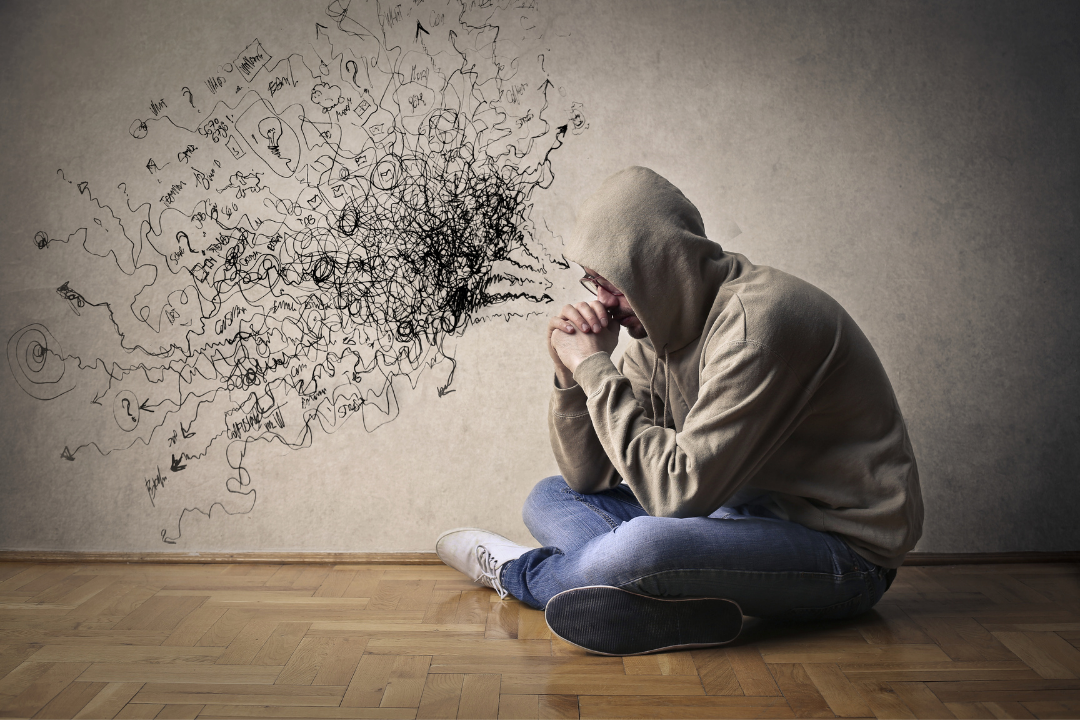 man in hoodie sitting on floor with negative thoughts spilling from head